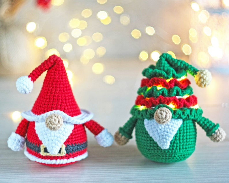 May include: Two crocheted gnomes, one in red and white and one in green and red, with white beards and light-up features. The gnomes are sitting on a wooden surface with a blurred background of white lights.