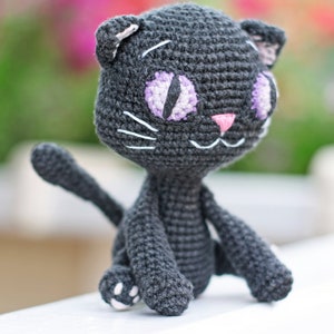 May include: A crocheted black cat toy with purple and white eyes and a pink nose. The cat is sitting on a white surface.