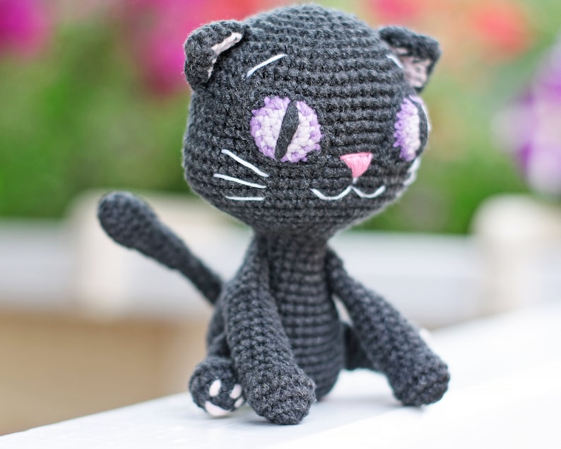 May include: A crocheted black cat toy with purple and white eyes and a pink nose. The cat is sitting on a white surface.