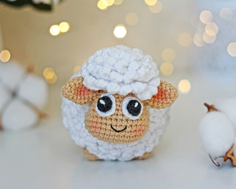 Pu&ograve; includere: Un agnello bianco all'uncinetto con tratti del viso marroni e rosa e occhi neri. L'agnello &egrave; seduto su una superficie bianca.