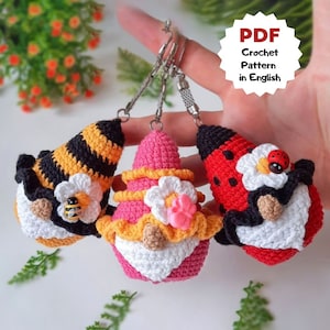 May include: Three crocheted gnome keychains with different designs. The first gnome is a bee with black and yellow stripes, the second gnome is pink with a white flower and a pink bow, and the third gnome is red with black spots and a white flower with a ladybug.