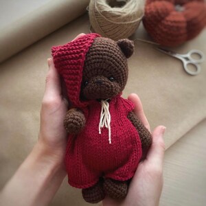 May include: A brown crocheted teddy bear wearing a red crocheted hooded jumpsuit. The bear is being held in the hands of a person.