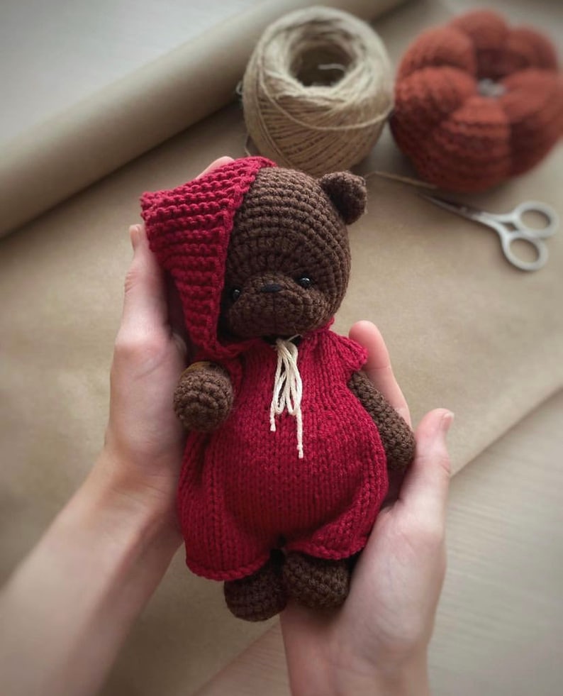 May include: A brown crocheted teddy bear wearing a red crocheted hooded jumpsuit. The bear is being held in the hands of a person.