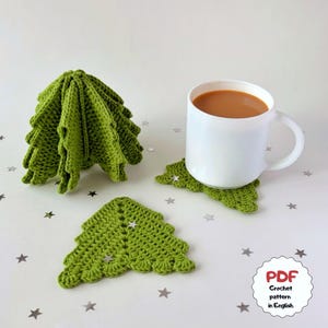 Puede incluir: Un conjunto de posavasos de ganchillo verde con forma de árbol, uno de ellos apilado para formar un árbol tridimensional, y una taza de cerámica blanca llena de líquido marrón. Una etiqueta blanca con texto rojo y negro dice PDF Crochet pattern in English.