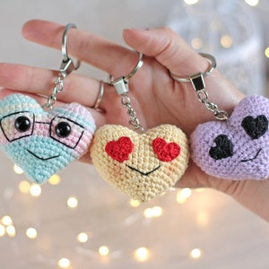 May include: Three crocheted heart-shaped keychains with different facial expressions. One has glasses, one has red heart eyes, and one has black heart eyes.