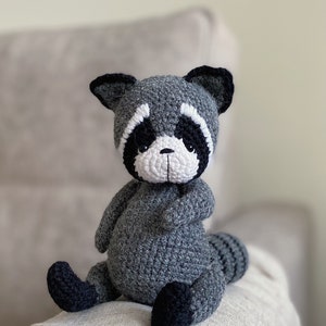 May include: A gray crocheted raccoon stuffed animal with black eyes, a white nose, and black ears. The raccoon is sitting on a light brown surface.