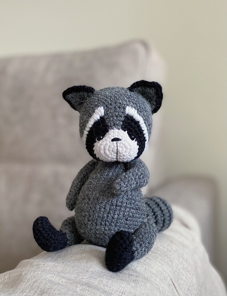 May include: A gray crocheted raccoon stuffed animal with black eyes, a white nose, and black ears. The raccoon is sitting on a light brown surface.