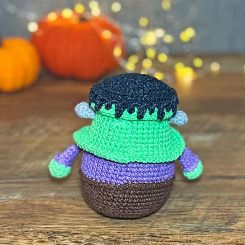 May include: A crocheted Frankenstein monster toy with green skin, purple pants, and a black hairdo. The toy is sitting on a wooden surface.