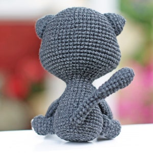 May include: A gray crocheted cat toy sitting on a white surface. The cat is facing away from the camera with its tail curled up.