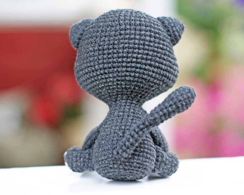 May include: A gray crocheted cat toy sitting on a white surface. The cat is facing away from the camera with its tail curled up.