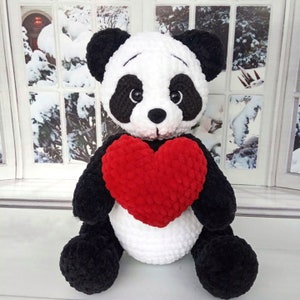 Peut inclure: Une peluche panda faite &agrave; la main, avec une fourrure noire et blanche, tenant un grand c&oelig;ur rouge. Le panda a des oreilles et des yeux noirs, et est assis devant une fen&ecirc;tre avec une sc&egrave;ne hivernale enneig&eacute;e.