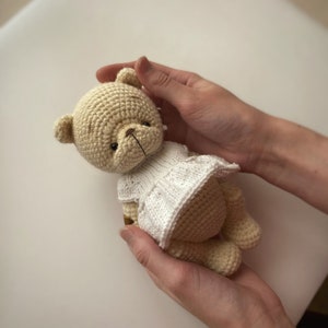 May include: A crocheted teddy bear wearing a white dress is held in two hands. The bear is light brown and has a brown nose and black eyes.