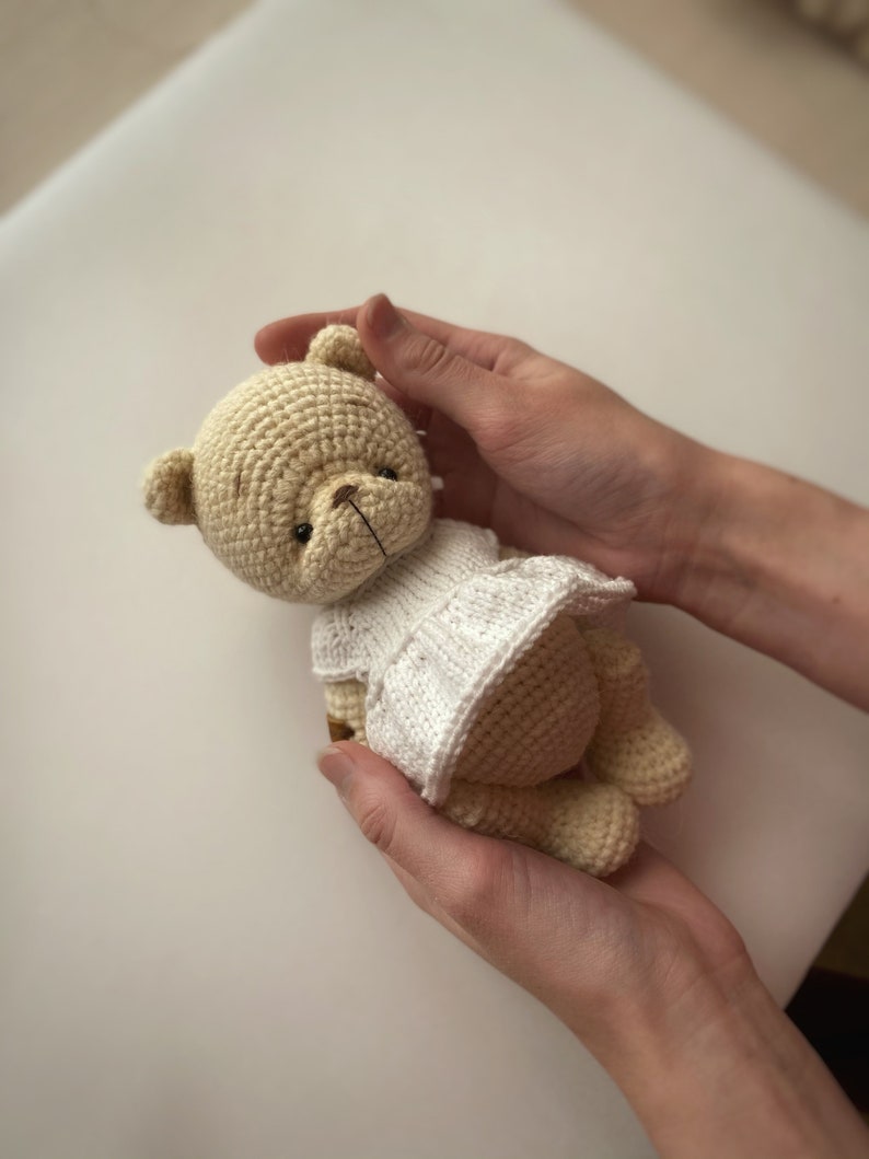 May include: A crocheted teddy bear wearing a white dress is held in two hands. The bear is light brown and has a brown nose and black eyes.