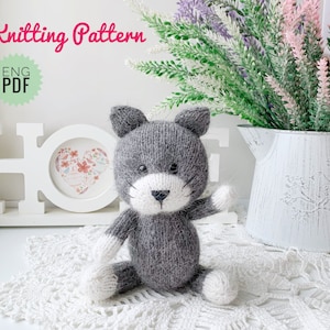 May include: Knitting pattern for a grey and white cat amigurumi toy. The pattern is available in English and is a PDF download. The cat is sitting on a white crocheted doily.