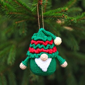 May include: A green crocheted gnome ornament with a red and white hat, a white beard, and a white pom-pom on top of the hat. The gnome is hanging from a green pine tree branch.