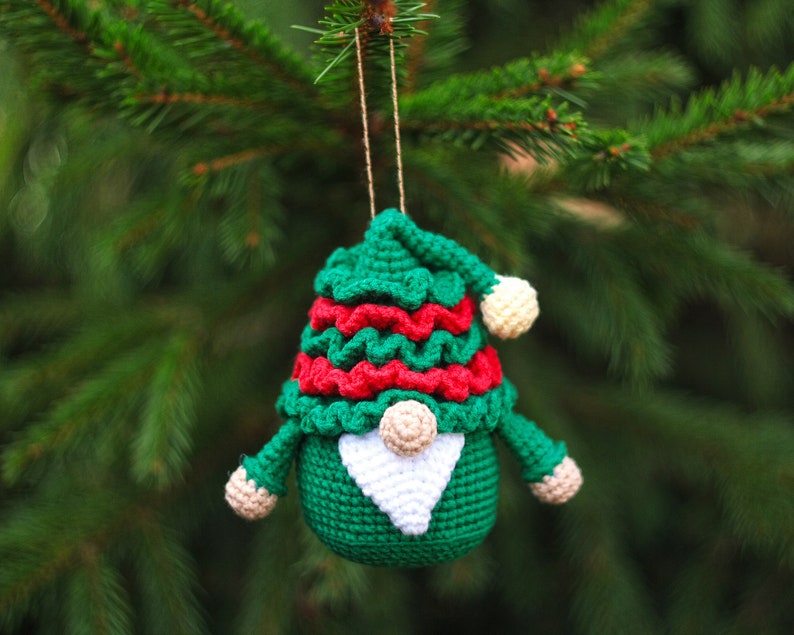 May include: A green crocheted gnome ornament with a red and white hat, a white beard, and a white pom-pom on top of the hat. The gnome is hanging from a green pine tree branch.