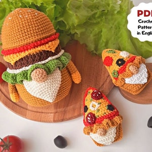 May include: Three crocheted gnome ornaments, one shaped like a hamburger, one shaped like a pizza slice, and one shaped like a pizza slice. The ornaments are on a wooden cutting board with lettuce, tomatoes, and olives.