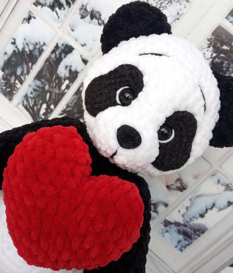 Peut inclure: Une peluche panda faite &agrave; la main, avec une fourrure noire et blanche, tenant un grand c&oelig;ur rouge. Le panda a des yeux noirs et un nez noir. Le jouet est fait d'une mati&egrave;re douce et textur&eacute;e.