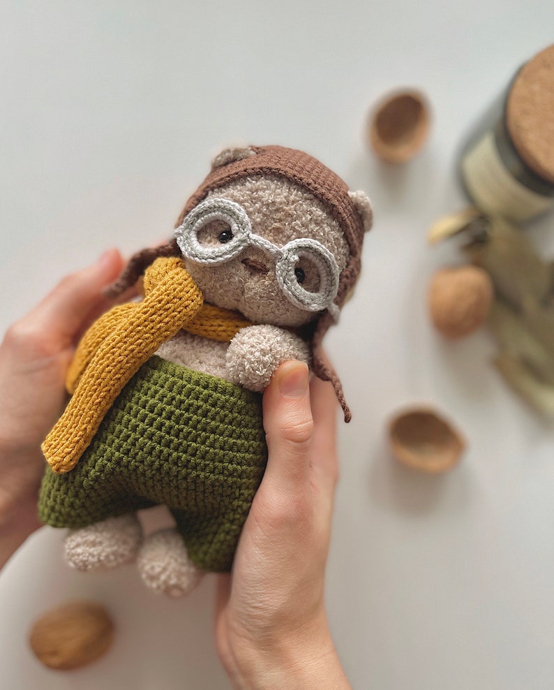 May include: A crocheted plush toy bear wearing a brown aviator hat, white glasses, and a yellow scarf. The bear is wearing green pants and is being held by hands.