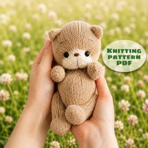 May include: A small, light brown knitted teddy bear toy with a cream-colored muzzle, dark round eyes, and a brown stitched nose. The toy features a soft, textured yarn construction and includes a circular graphic with the text KNITTING PATTERN PDF.