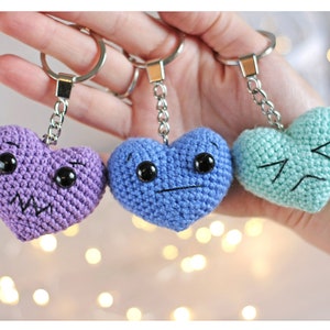 May include: Three crocheted heart-shaped keychains with different facial expressions. The hearts are purple, blue, and light green.