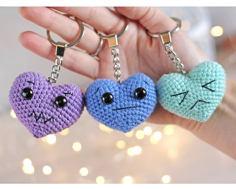 May include: Three crocheted heart-shaped keychains with different facial expressions. The hearts are purple, blue, and light green.