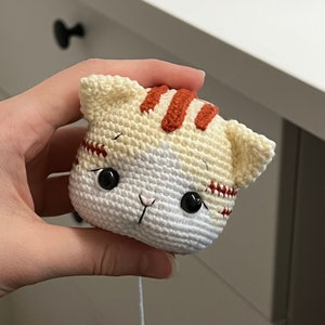 Puede incluir: Un juguete de crochet con forma de gato, hecho con hilo blanco y naranja. El gato tiene ojos negros y una nariz rosa.