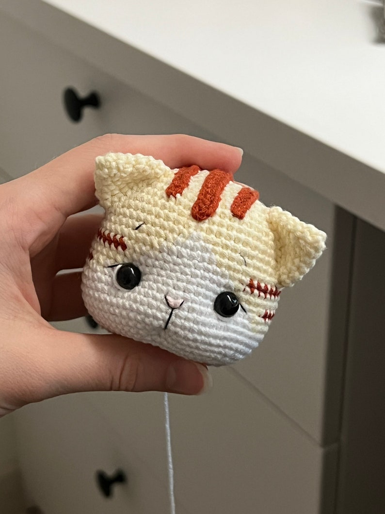 Puede incluir: Un juguete de crochet con forma de gato, hecho con hilo blanco y naranja. El gato tiene ojos negros y una nariz rosa.