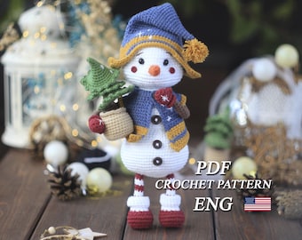 Könnte beinhalten: Ein gehäkelter Schneemann mit blauem Hut und Schal, der einen kleinen grünen Baum hält. Der Schneemann hat eine Karottennase, Knopfaugen und rote Fäustlinge. Der Text "PDF CROCHET PATTERN ENG" ist sichtbar. Die amerikanische Flagge ist ebenfalls sichtbar.