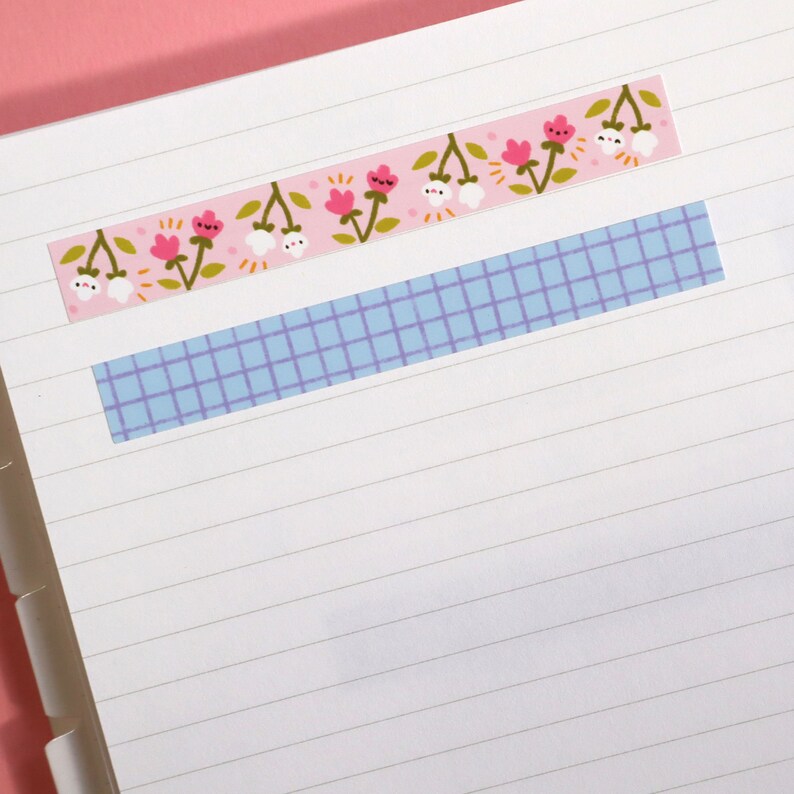 May include: Decorative washi tape strips on lined paper. One strip features a pink background with pink flowers and white floral accents. Another strip has a blue grid pattern. The paper is white with horizontal lines.