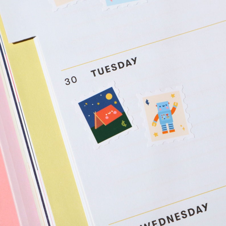 Pu&ograve; includere: Un'agenda bianca con la parola "TUESDAY" stampata in nero. L'agenda ha un adesivo di un robot blu e un adesivo di una tenda rossa con un cielo stellato.