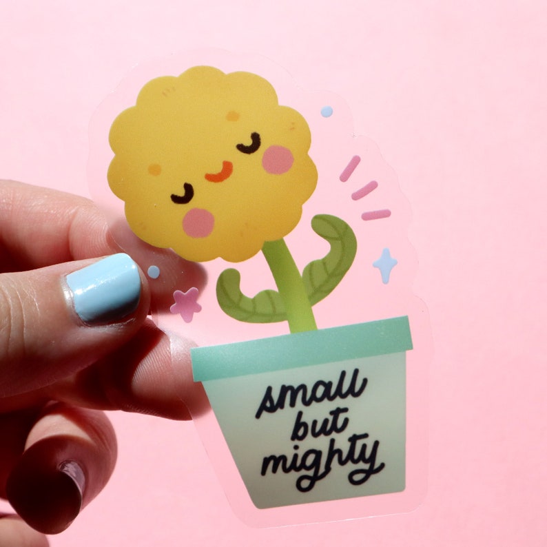 May include: A clear sticker with a yellow flower with a smiling face in a green pot. The pot has the text "small but mighty" written on it.