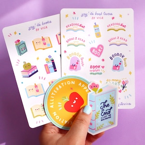 May include: A set of colourful book-themed stickers featuring illustrations of books, apples, ghosts, and a bookworm. The stickers are on a white background with a purple surface behind them. The stickers include the text "yay! it's books" and "yay! it's book covers" with the name "VICA" printed below. The stickers also include the text "reading list", "books I read", "booook lover", "book worm", and "so many books".