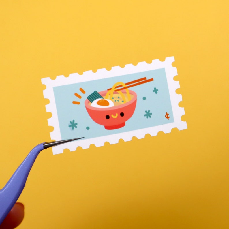 Op de afbeelding: Een witte en blauwe postzegel met een cartoonillustratie van een kom ramen met een glimlachend gezicht. De ramen bevat noedels, een groene zeewiergarnering en een wit ei. De kom is rood met een witte rand. De postzegel heeft een witte rand met een gegolfde rand.