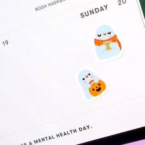 May include: Two cute ghost stickers with a white background. One ghost is holding a pumpkin and the other is holding a cup of coffee with a scarf around its neck.