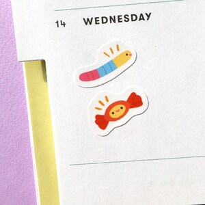May include: Two colourful stickers on a white background. The first sticker is a red and blue worm with a yellow head and a smiling face. The second sticker is a red and white candy with a yellow head and a smiling face.
