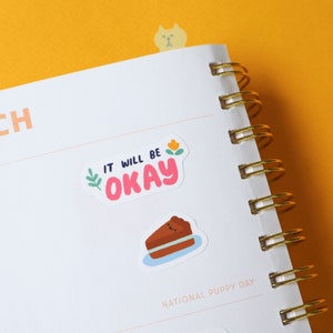 May include: A white planner with a gold spiral binding. The planner has two stickers on it. One sticker says "It will be okay" with a pink background and a yellow flower. The other sticker is a brown slice of cake with a happy face.