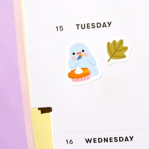 May include: A blue ghost wearing a red hat is holding a knife and decorating a pumpkin pie. A brown leaf is in the background. The text "TUESDAY" and "15" are visible.