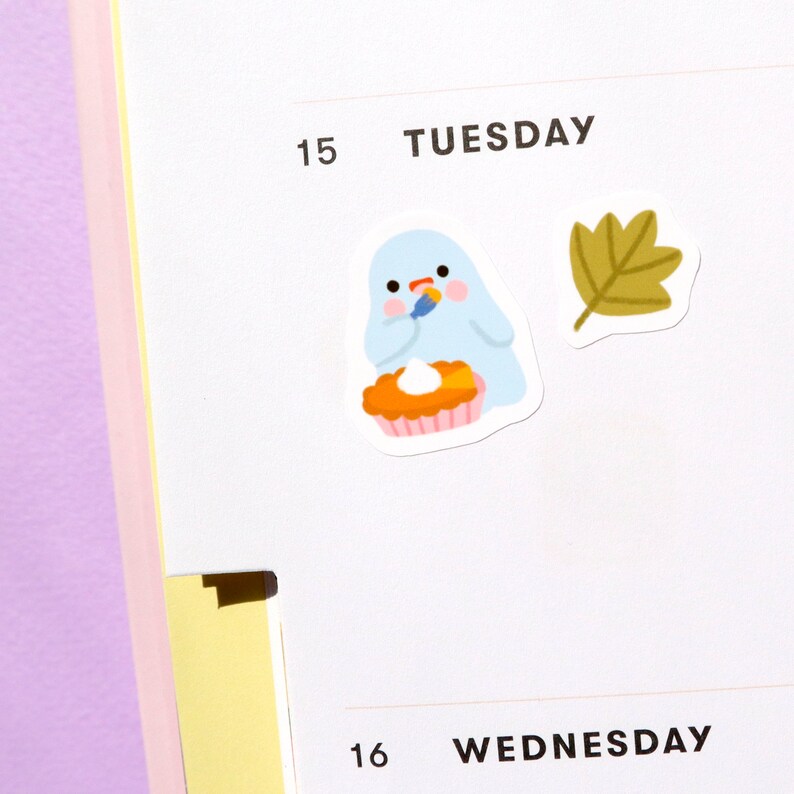 May include: A blue ghost wearing a red hat is holding a knife and decorating a pumpkin pie. A brown leaf is in the background. The text "TUESDAY" and "15" are visible.