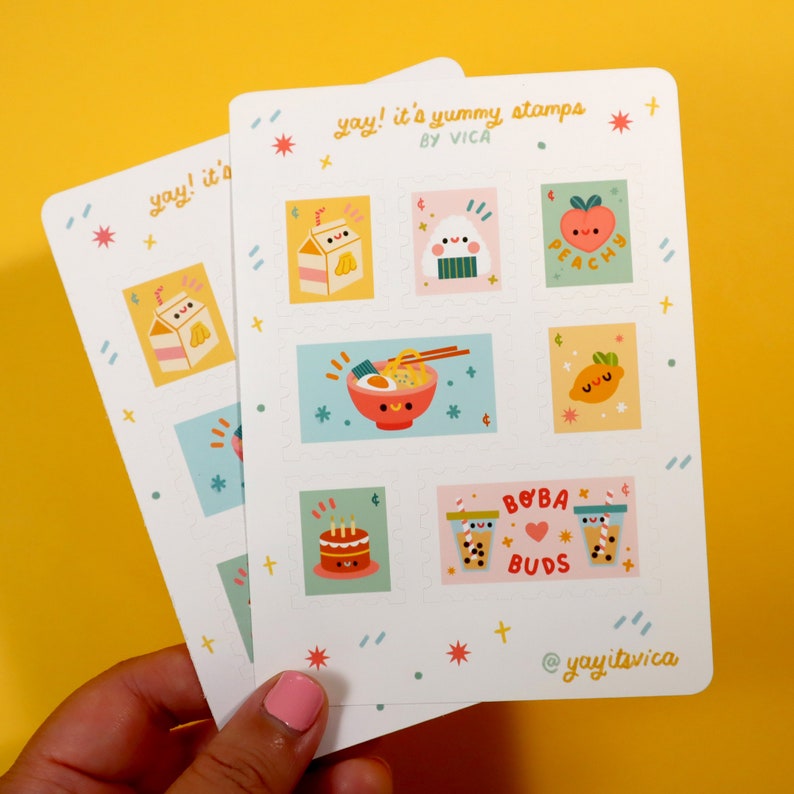 Op de afbeelding: Een vel met kleurrijke stickers met schattige illustraties van eten en drinken. De stickers hebben de vorm van postzegels en bevatten ontwerpen zoals een melkpak, een rijstbal, een perzik, een kom ramen, een citroen en een boba-thee. De tekst op het vel luidt "yay! it's yummy stamps by VICA" en "BOBA BUDS".