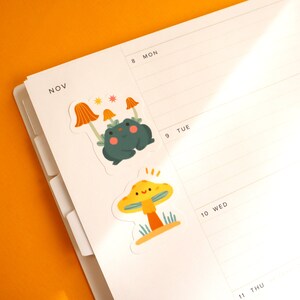 May include: Two colorful stickers featuring a green frog with a mushroom cap and a yellow mushroom with a smiling face. The stickers are placed on a white planner with the month of November and days of the week labeled.
