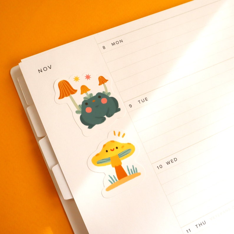 May include: Two colorful stickers featuring a green frog with a mushroom cap and a yellow mushroom with a smiling face. The stickers are placed on a white planner with the month of November and days of the week labeled.