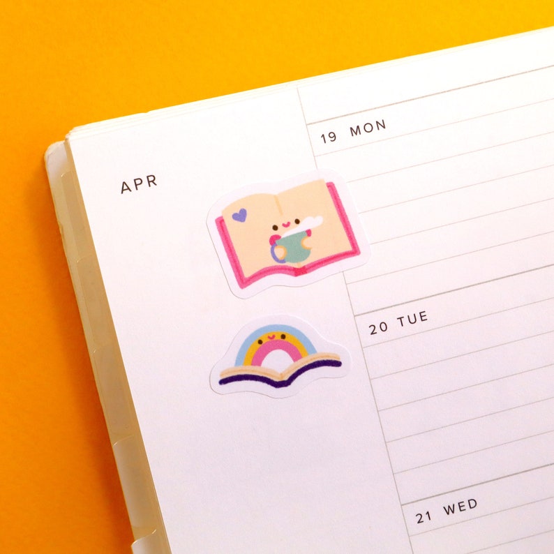 May include: Two colourful stickers featuring a book with a rainbow and a book with a cup of coffee. The stickers are on a white planner with the month of April and the days of the week written on it.