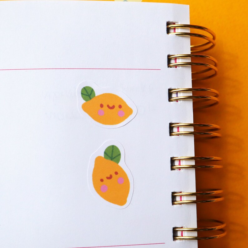 Puede incluir: Dos pegatinas de lim&oacute;n amarillo con una cara sonriente y una hoja verde en un cuaderno blanco con anillos dorados.