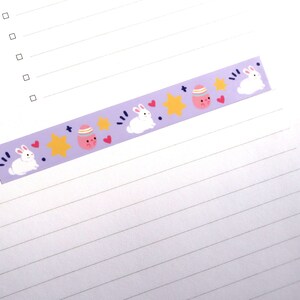 May include: A decorative purple washi tape featuring Easter-themed illustrations. The tape showcases white bunnies, pink eggs with faces, yellow stars, and red hearts. The tape is designed for crafts and scrapbooking.