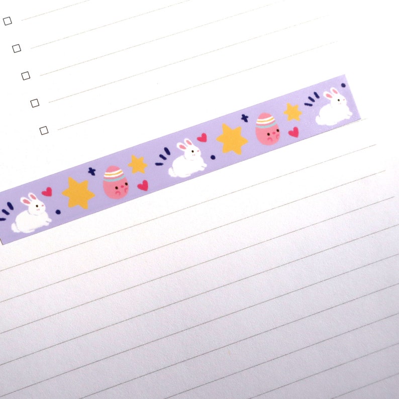 May include: A decorative purple washi tape featuring Easter-themed illustrations. The tape showcases white bunnies, pink eggs with faces, yellow stars, and red hearts. The tape is designed for crafts and scrapbooking.