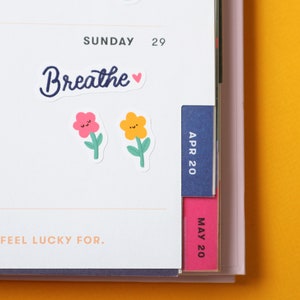 May include: A white planner with a blue tab labeled "APR 20" and a pink tab labeled "MAY 20". The planner has a sticker that says "Breathe" with a heart. There are also two flower stickers with smiley faces.