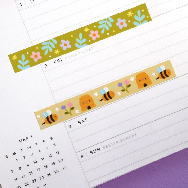 May include: Two decorative washi tape strips with floral and bee designs. One strip is olive green with pink and purple flowers and blue leaves. The other is tan with bees, beehives, and flowers. The tape is on a calendar page.