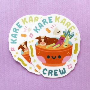 K&ouml;nnte beinhalten: Zwei wei&szlig;e Aufkleber mit einer Cartoon-Illustration einer Sch&uuml;ssel Suppe mit einer Kuh, Aubergine, Knoblauch und Fr&uuml;hlingszwiebeln. Der Text "KARE KARE CREW" ist um die Sch&uuml;ssel herum geschrieben.