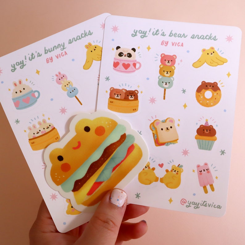 Może przedstawiać: Dwa zestawy naklejek z uroczymi ilustracjami zwierząt. Pierwszy zestaw nosi tytuł "yay! it's bunny snacks" i przedstawia kr&oacute;liki, hamburgera i inne przekąski. Drugi zestaw nosi tytuł "yay! it's bear snacks" i przedstawia niedźwiedzie, pączka i inne przekąski.